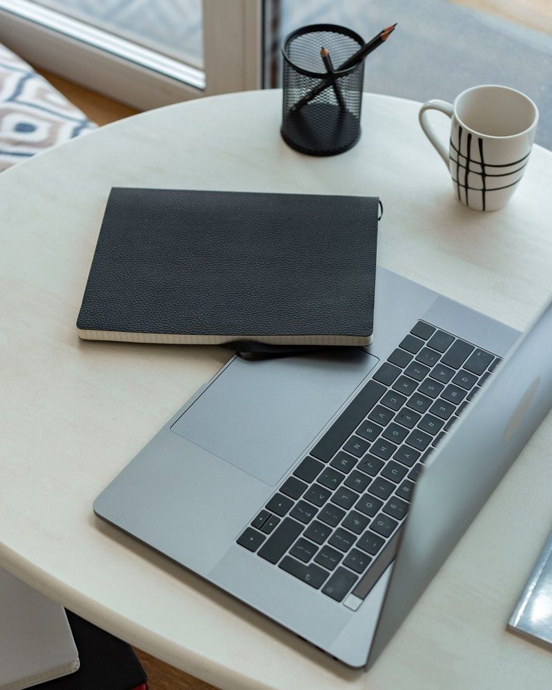 A laptop, notebook and mug on a desk by a window
